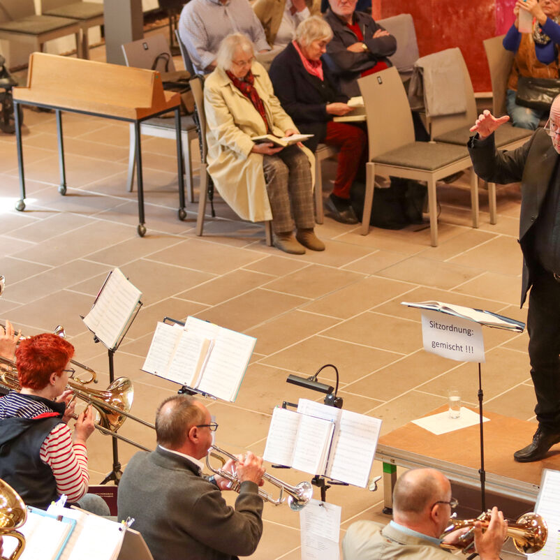Ein schwarzgekleideter Dirigent steht vor Musizierenden mit Notenständern.