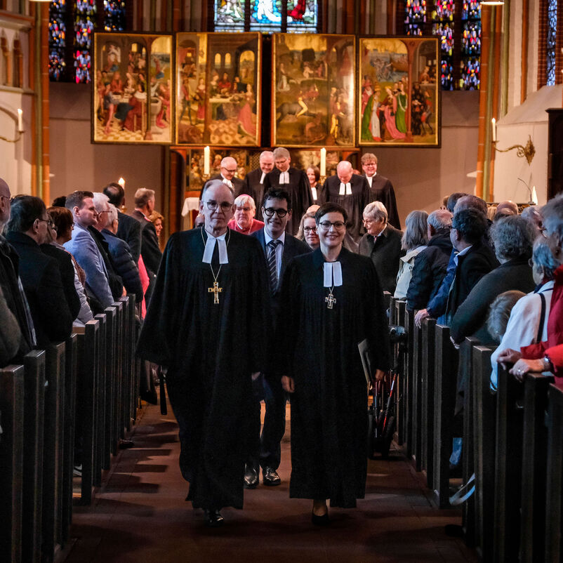 Eine männliche und eine weiblich zu lesende Person laufen in Talaren einen Mittelgang in einer Kirche entlang, sie führen eine Delegation an. Links und rechts stehen viele Menschen. Im Hintergrund leuchtet ein goldenes Altarbild.