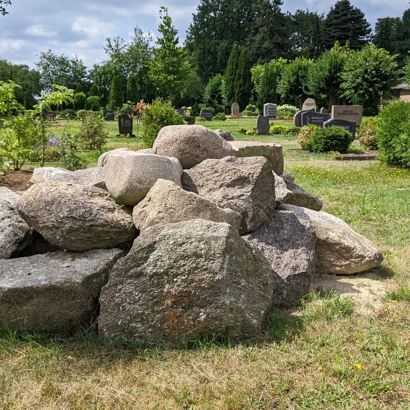 Steine sind zu einem großen Haufen geschichtet, im Hintergrund sind Gräber eines Friedhofs zu erkennen.