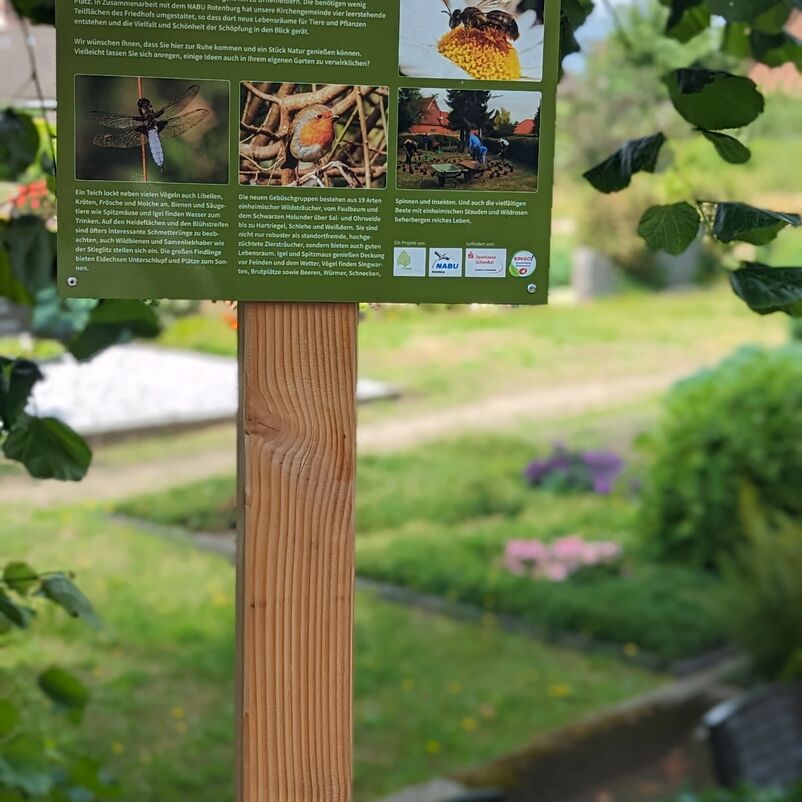 An einem Holzpfahl ist ein Schild montiert, das auf die Umweltschutzmaßnahmen auf dem Friedhof hinweist.