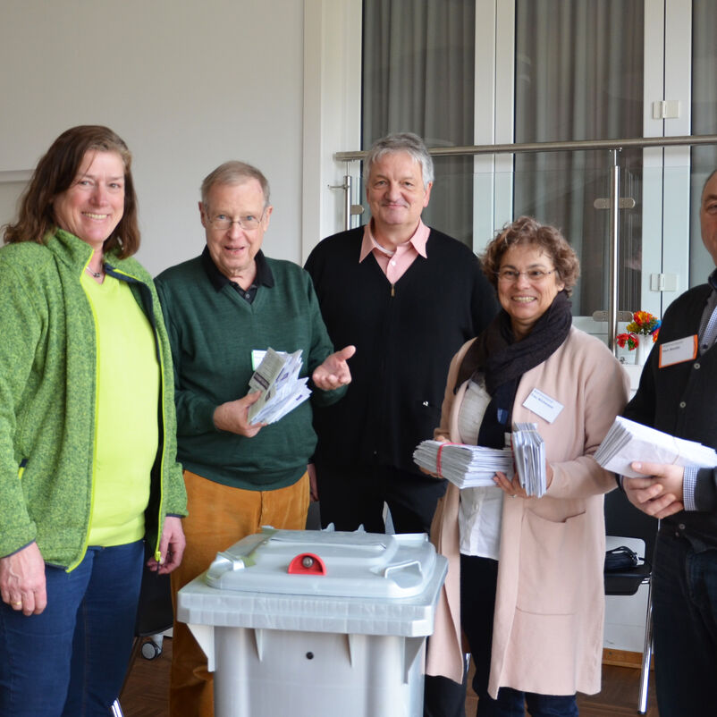 Fünf Personen stehen um eine graue Wahlurne und haben Zettel in Händen.