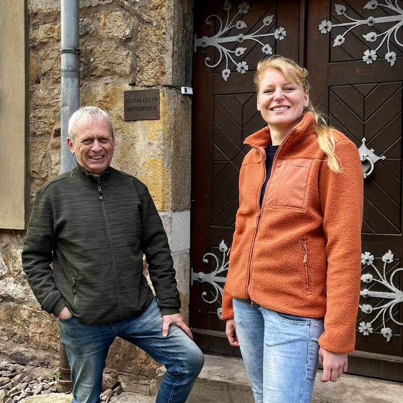 Eine weiblich gelesene Person mit orangefarbener Jacke und langen blonden Haaren steht mit einer männlich gelesenen Person in Outdoor-Jacke vor einer Haustür mit dem Schild "Kloster Loccum Forstverwaltung".