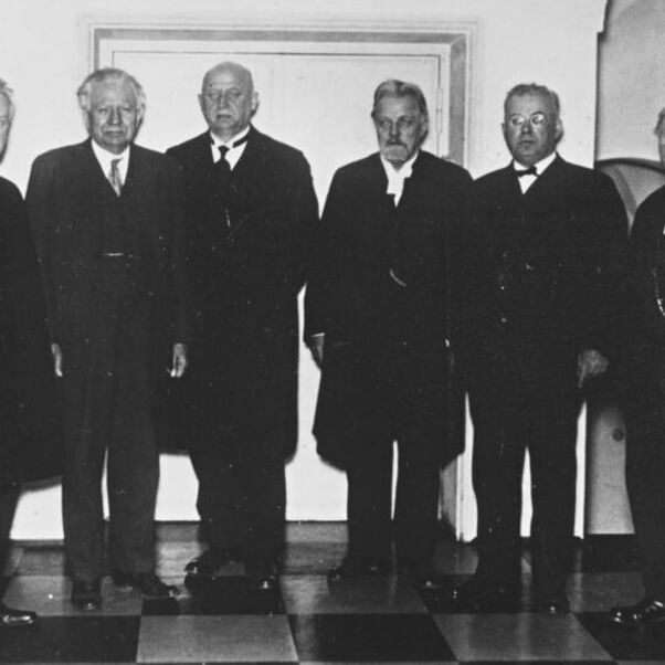 Das Schwarzweiß-Bild zeigt sechs männlich lesbare Personen in schwarzen Anzügen stehend für das Foto vor einer weißen Wand.