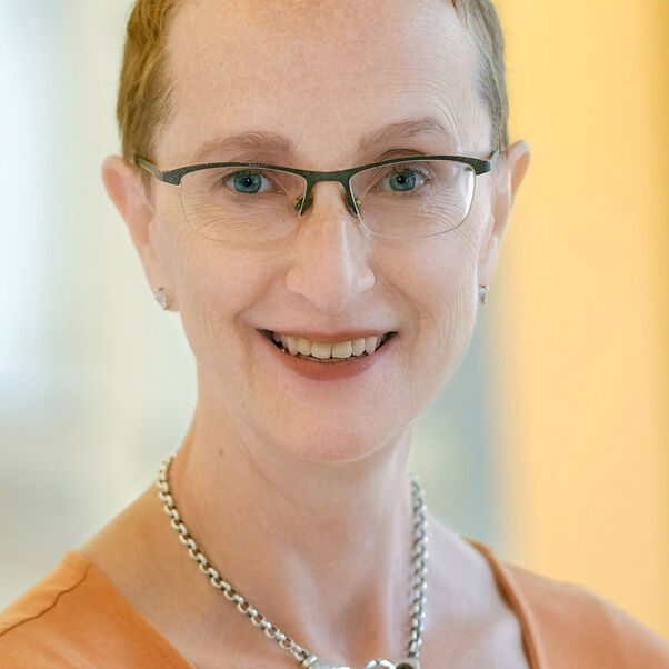 Das Bild zeigt eine weiblich lesbare Person mit sehr kurzen rotbraunen Haaren, einer Brille mit Oberfassung, einem orangefarbenen Oberteil und einer silbernen Kette.