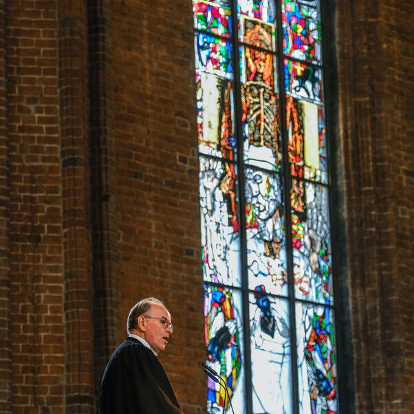 Eine männlich lesbare Person im schwarzen Talar auf einer Kanzel vor einem Kirchenfenster.