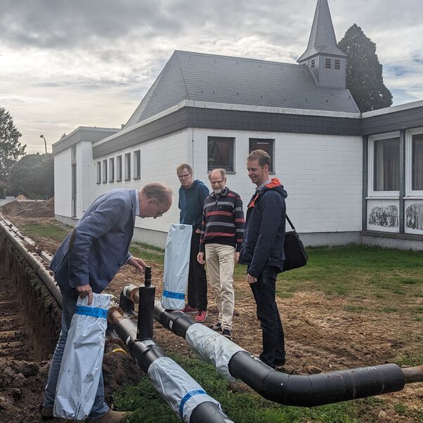 Vier Menschen schauen sich schwarze Rohre an, die neben einem Graben liegen. Im Hintergrund ist ein Haus und ein Kirchengebäude zu sehen.